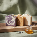 Three goat milk soap bars arranged on weathered oak board — lavender, oatmeal honey, and unscented
