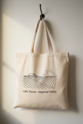 Natural cotton AMC Farms tote bag hanging on a hook with farm field illustration